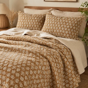 Pode incluir: Um conjunto de cama composto por uma colcha e duas fronhas num tom castanho-mostarda quente com um padrão floral branco repetitivo. O conjunto está sobreposto a uma manta texturizada de cor creme sólida para a cama.