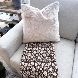 May include: A plush, light beige faux fur pillow rests on a gray sofa, paired with a brown and white patterned throw blanket. The throw features a repeating leaf design. The scene suggests a cozy living room setting.