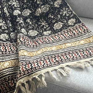 May include: A black and cream floral patterned textile with a cream fringe. The textile has a repeating floral design and a border with geometric patterns in cream, brown, and red.