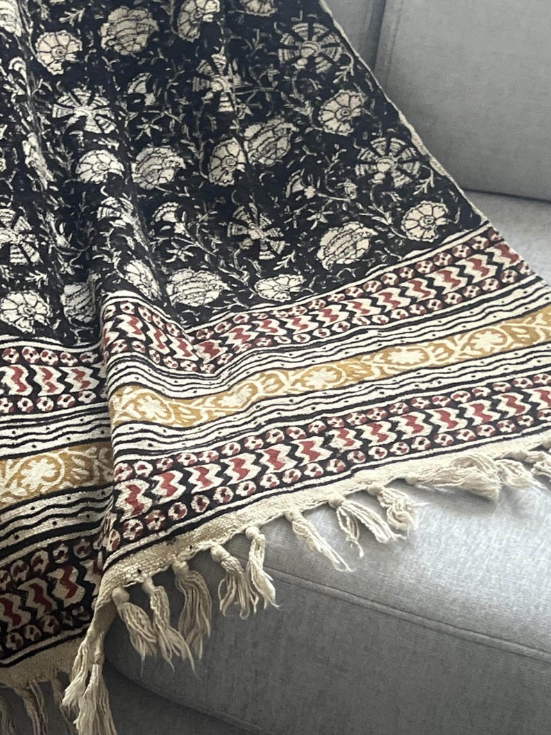 May include: A black and cream floral patterned textile with a cream fringe. The textile has a repeating floral design and a border with geometric patterns in cream, brown, and red.