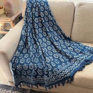 May include: A deep blue throw blanket featuring a repeating white floral block print pattern. The fabric has a textured appearance and is finished with a decorative zigzag border and knotted fringe along the bottom edge.