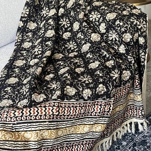 May include: Black and cream floral patterned throw blanket with a decorative border featuring geometric and floral designs in red, cream, and gold. The blanket has a fringed edge and is draped over a couch.