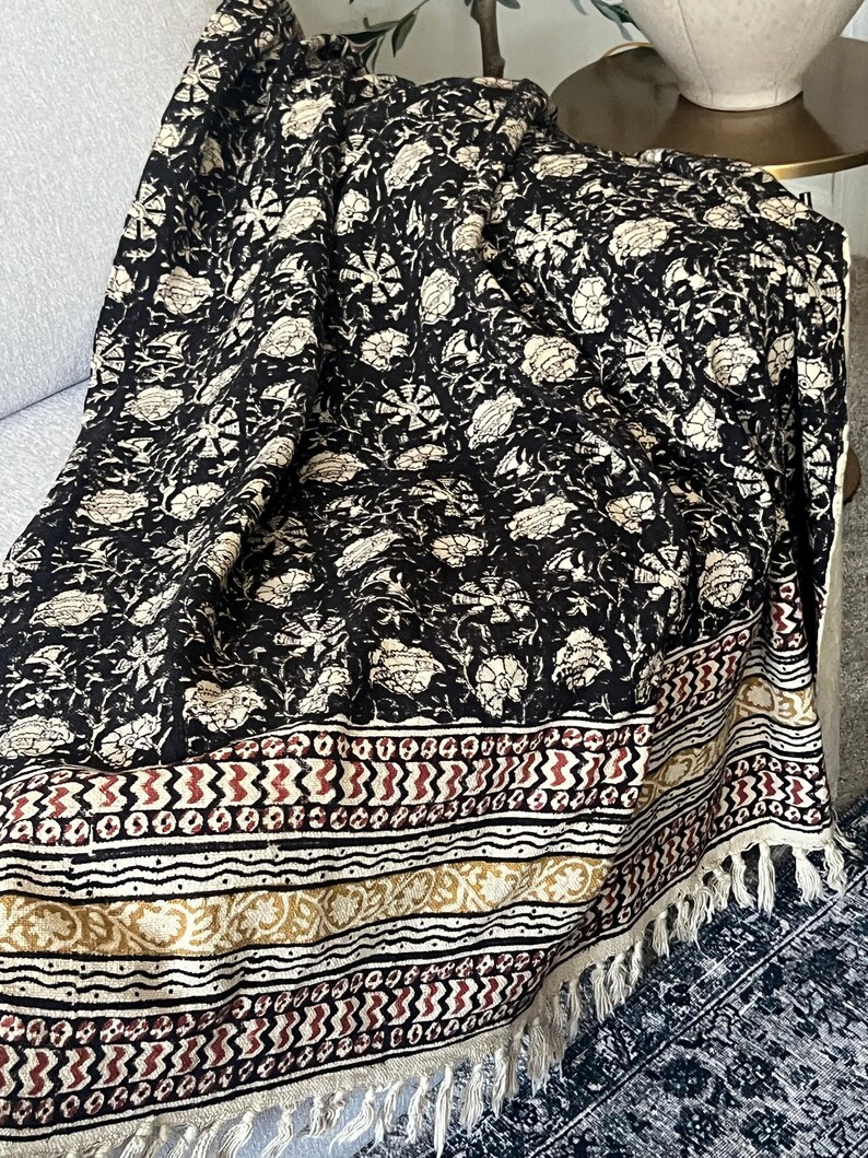 May include: Black and cream floral patterned throw blanket with a decorative border featuring geometric and floral designs in red, cream, and gold. The blanket has a fringed edge and is draped over a couch.