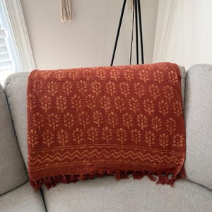 May include: A rectangular throw blanket in a deep rust-orange color featuring a repeating pattern of small, light-colored floral motifs. The bottom edge is finished with a horizontal zigzag band and a row of matching rust-colored fringe tassels.
