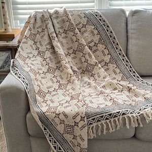 May include: A beige throw blanket with a geometric pattern in brown and black is draped over a gray sofa. The blanket features a repeating diamond and zigzag design, with a fringed edge. The sofa is positioned in front of a window with white blinds.