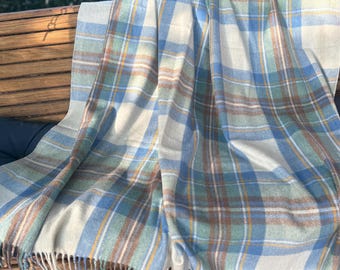 May include: A woven throw blanket featuring a plaid pattern in shades of light blue, sage green, cream, and brown. The fabric has a soft texture with visible weave lines and a fringe trim along the bottom edge, creating a classic textile design.