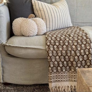 May include: A cozy living room scene featuring a light gray sofa adorned with decorative pillows and a patterned throw blanket. A plush, cream-colored pumpkin adds a touch of autumn. The throw blanket has a brown floral pattern.