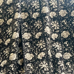 May include: Black fabric with a repeating floral pattern in cream and off-white. The design features large, stylized flowers and smaller floral elements. The fabric has a textured appearance, with visible folds and drapes. A decorative border with red and brown accents is visible.
