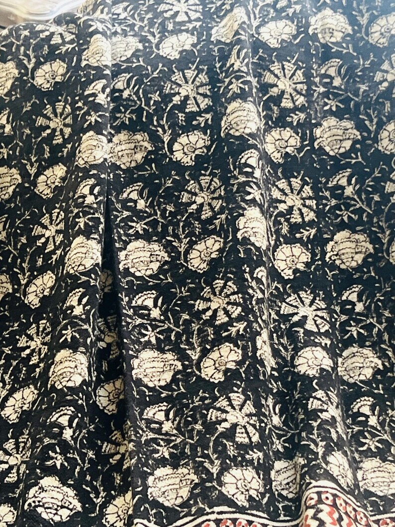 May include: Black fabric with a repeating floral pattern in cream and off-white. The design features large, stylized flowers and smaller floral elements. The fabric has a textured appearance, with visible folds and drapes. A decorative border with red and brown accents is visible.