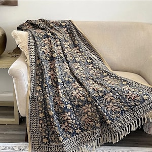 May include: A rectangular throw blanket featuring a dark blue background with a repeating floral pattern in shades of tan, cream, and muted red. The blanket has a wide decorative border with a black and white zigzag design and cream fringe.