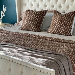 Peut inclure: Un lit avec une tête de lit capitonnée blanche à clous argentés, deux coussins carrés marron à motif floral blanc, un couvre-lit floral marron assorti et un jeté de lit à chevrons gris posé au pied du lit.