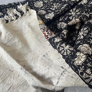 May include: A folded textile with a cream-colored section and a black section. The black section features a floral pattern in cream. The cream section has a fringed edge and a patterned border with red and brown accents.