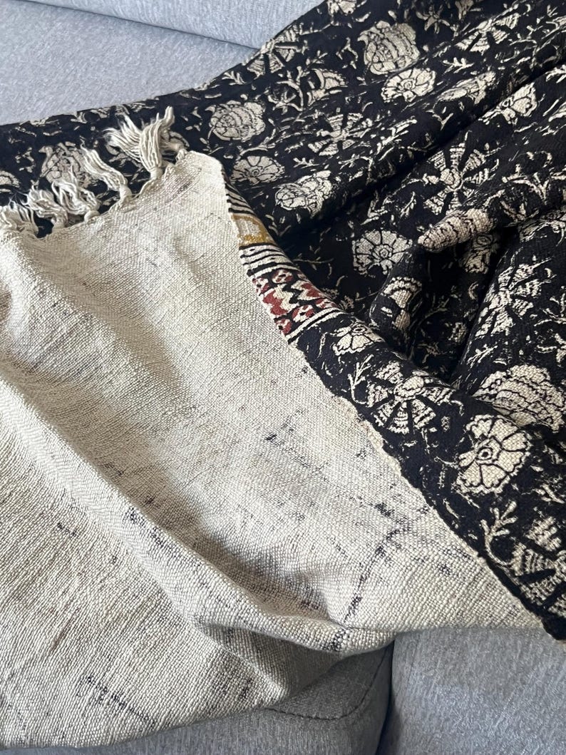 May include: A folded textile with a cream-colored section and a black section. The black section features a floral pattern in cream. The cream section has a fringed edge and a patterned border with red and brown accents.
