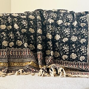 May include: A black and cream floral patterned throw blanket with a decorative border in burgundy and gold. The blanket has cream-colored tassels along the bottom edge and is draped over a gray sofa.