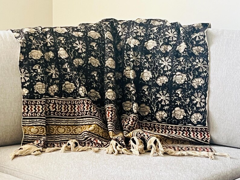 May include: A black and cream floral patterned throw blanket with a decorative border in burgundy and gold. The blanket has cream-colored tassels along the bottom edge and is draped over a gray sofa.
