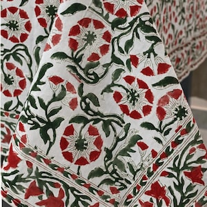Op de afbeelding: Een wit tafelkleed met een herhalend bloemenpatroon in donkerrood en donkergroen. Het ontwerp bevat gestileerde bloesems en bladerrijke ranken, met een duidelijk randpatroon langs de zijkanten van de vallende stof.