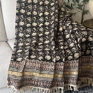 May include: A black and cream floral patterned throw blanket with a fringed edge. The blanket is draped over a light-colored sofa. The design includes floral and geometric patterns in cream, red, and gold.