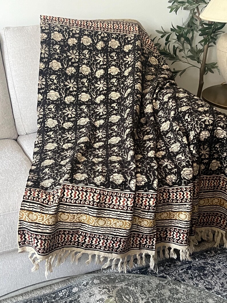 May include: A black and cream floral patterned throw blanket with a fringed edge. The blanket is draped over a light-colored sofa. The design includes floral and geometric patterns in cream, red, and gold.