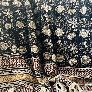 May include: A black and white floral patterned textile with a cream-colored fringe. The fabric features a repeating floral design in white against a black background. The border has a cream, red, and gold geometric pattern.