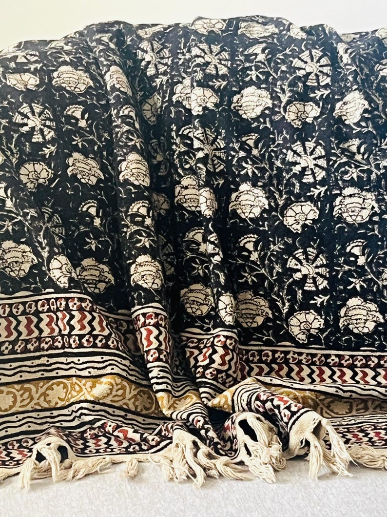 May include: A black and white floral patterned textile with a cream-colored fringe. The fabric features a repeating floral design in white against a black background. The border has a cream, red, and gold geometric pattern.