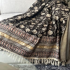 May include: A black and cream floral patterned throw blanket with a decorative border in cream, red, and gold. The blanket has a fringe trim and is draped over a gray sofa.
