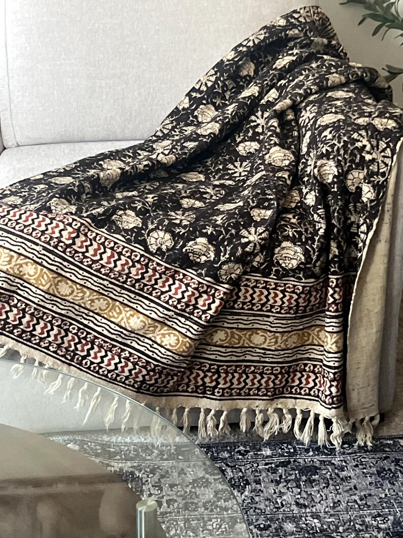 May include: A black and cream floral patterned throw blanket with a decorative border in cream, red, and gold. The blanket has a fringe trim and is draped over a gray sofa.