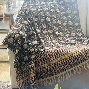 May include: A dark brown throw blanket with a floral pattern in cream and beige, draped over a light-colored sofa. The blanket has a decorative border with red, gold, and cream accents, and a fringed edge.