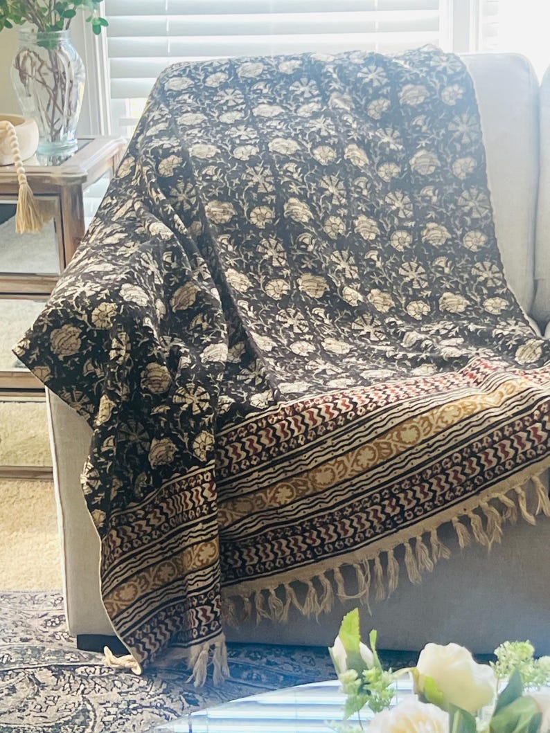 May include: A dark brown throw blanket with a floral pattern in cream and beige, draped over a light-colored sofa. The blanket has a decorative border with red, gold, and cream accents, and a fringed edge.