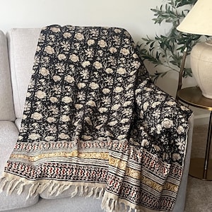 May include: A black and cream floral patterned throw blanket with a fringed edge is draped over a gray sofa. The blanket features a repeating floral design and a decorative border with geometric patterns in cream, red, and gold.