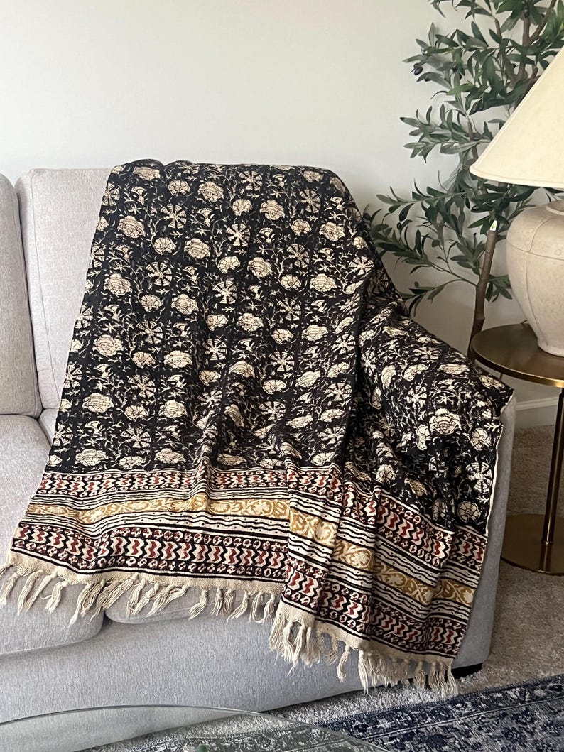 May include: A black and cream floral patterned throw blanket with a fringed edge is draped over a gray sofa. The blanket features a repeating floral design and a decorative border with geometric patterns in cream, red, and gold.