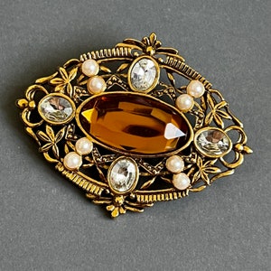 May include: A gold-toned brooch with a large, oval-shaped amber-colored stone surrounded by smaller clear stones and white pearls. The brooch is set in a delicate, intricate design.