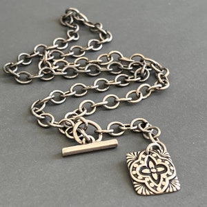 May include: A silver chain necklace with a square pendant featuring a detailed floral design. The necklace has a toggle clasp.