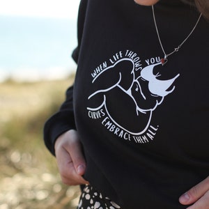 May include: Black sweatshirt with a white graphic design that says "When life throws you curves, embrace them all." The graphic features a stylized image of a person's arms and torso. The person is wearing a necklace with a small red heart pendant.