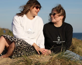 May include: Two people wearing sweatshirts with text on them are sitting on a grassy dune. The woman on the left is wearing a white sweatshirt with the text "If the weather can be so far two days and still feel colder can I"
