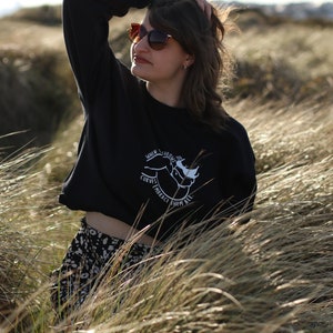 May include: A woman wearing a black cropped sweatshirt with a white graphic that says "When the curves embrace them all". She is wearing a black and white patterned skirt and sunglasses.