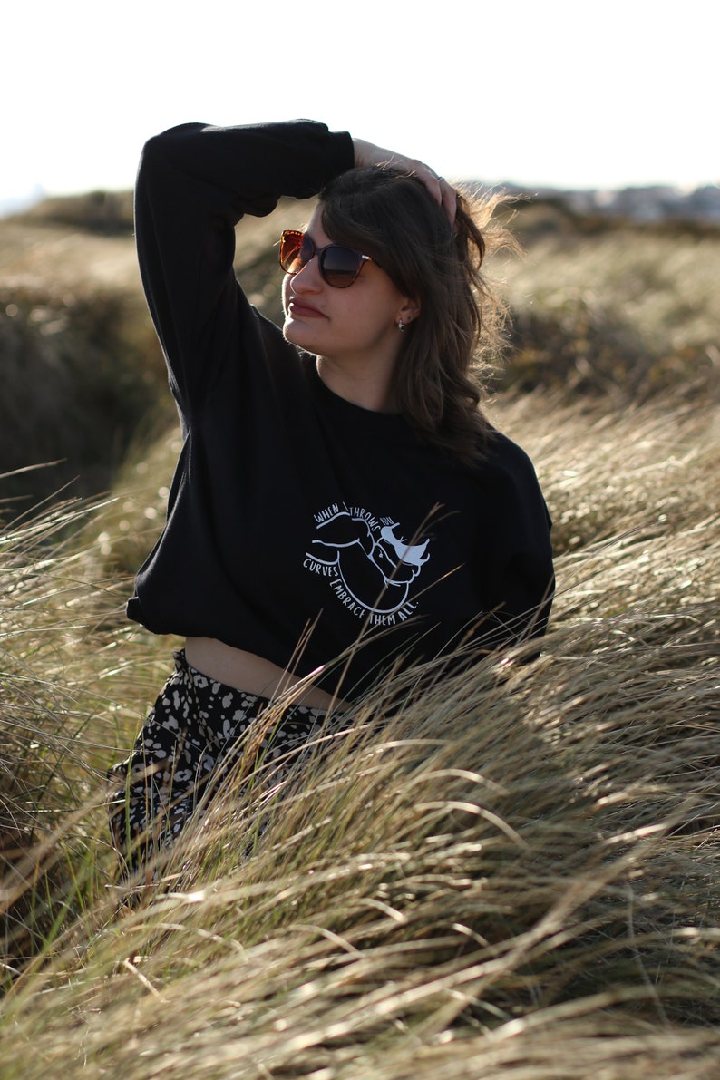 May include: A woman wearing a black cropped sweatshirt with a white graphic that says "When the curves embrace them all". She is wearing a black and white patterned skirt and sunglasses.