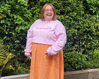 May include: A woman wearing a pink sweatshirt with the text "love got my girls" embroidered on the front, and a bright orange gingham skirt.