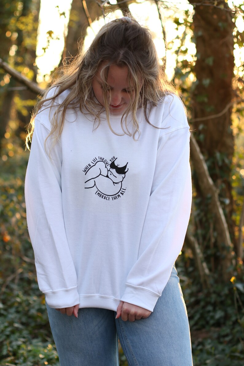 May include: White long-sleeve sweatshirt with a black graphic print of a hand holding a heart with the text "When life throws you, embrace them all."