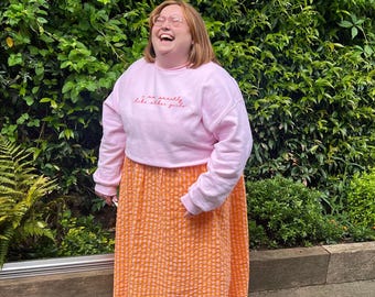 May include: A woman wearing a pink sweatshirt with the text "I'm actually like other girls" in red. She is wearing a bright orange and white checkered skirt and laughing.
