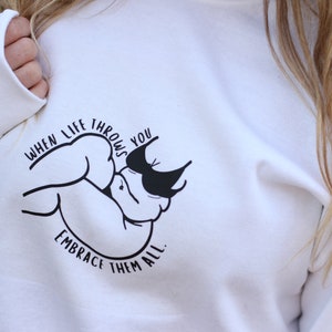 May include: White sweatshirt with a black graphic design that says "When life throws you, embrace them all." The graphic is a stylized image of a person catching a ball.