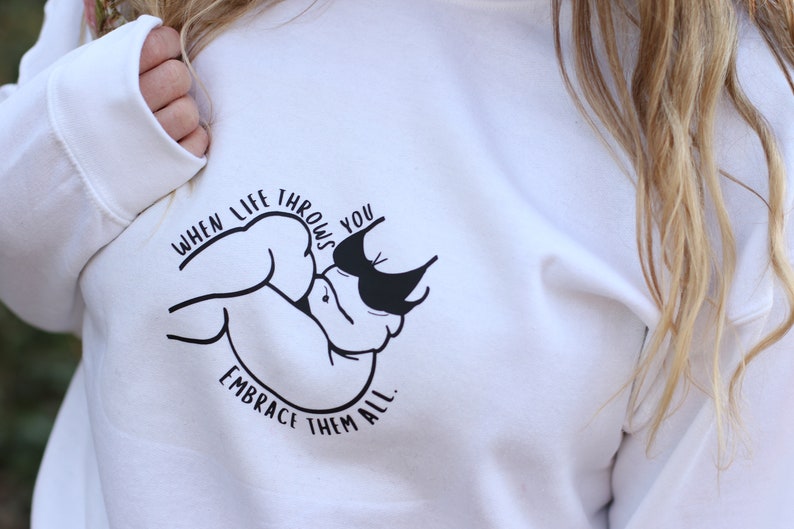 May include: White sweatshirt with a black graphic design that says "When life throws you, embrace them all." The graphic is a stylized image of a person catching a ball.