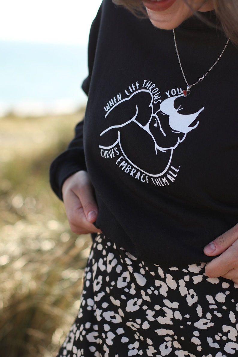 May include: Black sweatshirt with a white graphic design that says "When life throws you curves, embrace them all." The graphic features a stylized image of a person's arms and torso. The person is wearing a necklace with a small red heart pendant.