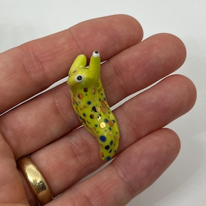 May include: A small, green ceramic slug with gold and blue spots. The slug has two white eyes and is posed in a curled position.
