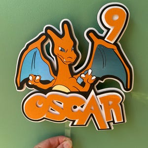 May include: A layered orange and blue Charizard cake topper with the number 9 and the name "OSCAR". The Charizard has blue wings and is set against a green background. The topper is held by a clear plastic stick.