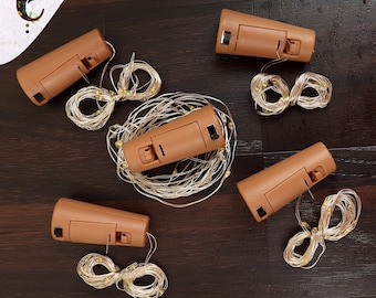 May include: Several brown cork-shaped LED string lights, each with a battery compartment and a string of small, warm white lights. The lights are arranged on a dark brown wooden surface.