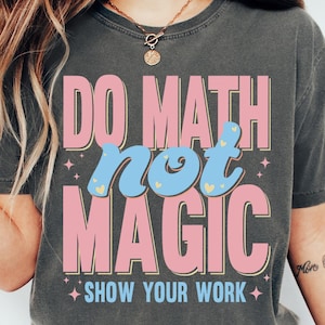 May include: A dark gray t-shirt with the text "DO MATH NOT MAGIC" in pink and blue, with the phrase "SHOW YOUR WORK" below. The shirt is a casual style, suitable for everyday wear.