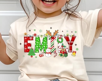 May include: A child wearing a cream coloured t-shirt with the name "EMILY" spelled out in colourful letters with a Christmas theme. The letters are decorated with Christmas trees, ornaments, candy canes, and other festive elements.