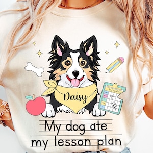 May include: A white t-shirt with a cartoon image of a dog wearing a yellow bandana with the name "Daisy" on it. The text "My dog ate my lesson plan" is printed below the dog. The shirt is available in ivory.