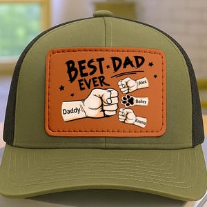 May include: Olive green and black trucker hat with a brown leather patch. The patch reads "BEST DAD EVER" with fist and paw illustrations, and the names "Daddy", "Alex", "Bailey", and "Emma".