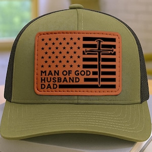 May include: An olive green and black trucker hat with a rectangular patch featuring an American flag design and a cross. The patch includes the text "MAN OF GOD HUSBAND DAD". The hat is sitting on a stack of books.
